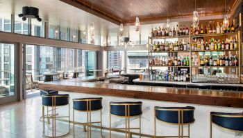 A modern bar with a variety of bottles on display, high stools, and an expansive view of a cityscape through large windows.