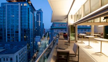 A modern cityscape with tall buildings at dusk and an elegant balcony with seating and tables, illuminated by warm lighting.