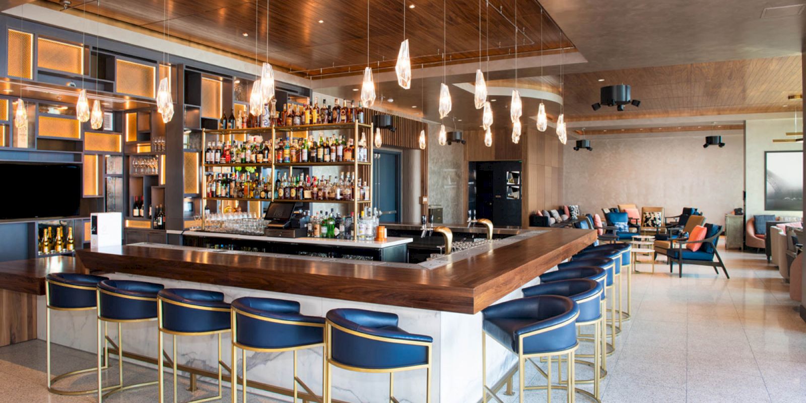 The image shows a modern bar with a central counter, blue bar stools, an extensive display of bottles, elegant lighting, and seating in the background.