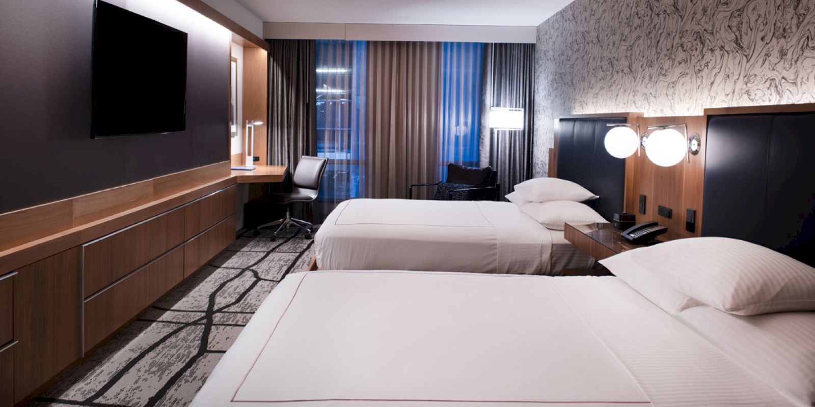 A modern hotel room with two beds, a flat-screen TV, a desk with a chair, and large windows with drapes.