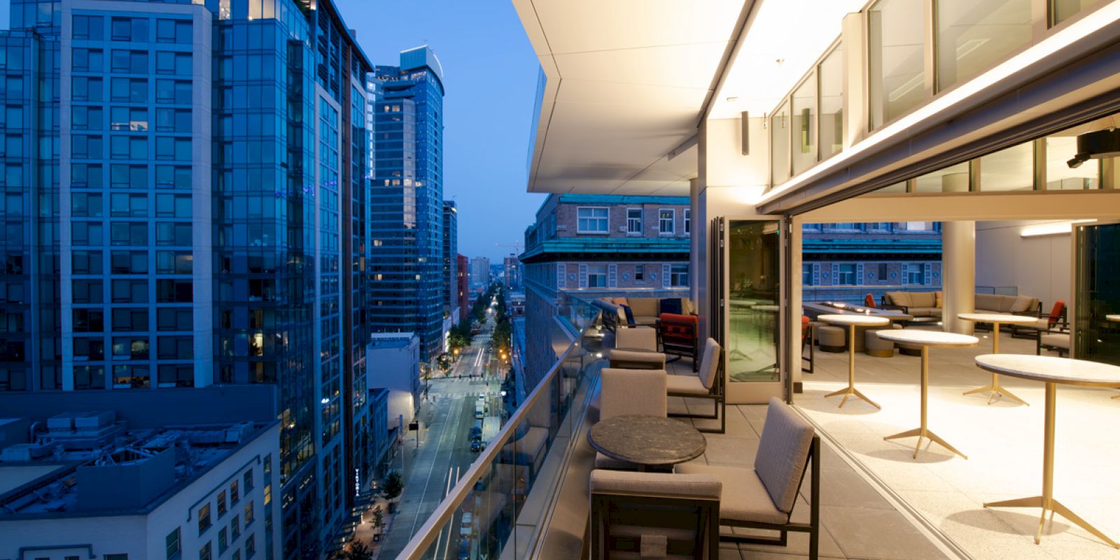 A modern rooftop terrace at dusk overlooking a cityscape with high-rise buildings and street lights. The terrace has seating and tables.