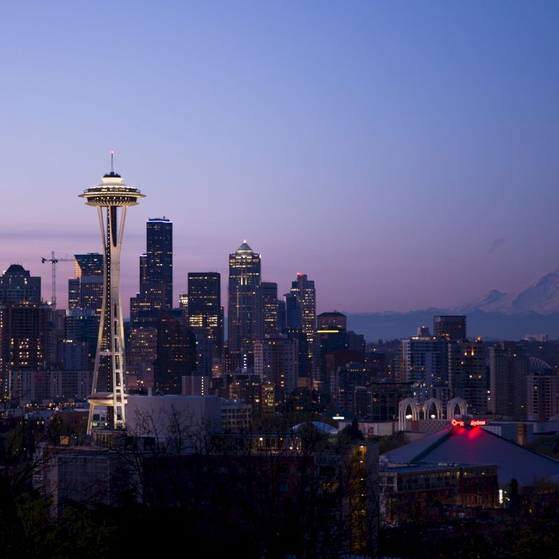 A panoramic view of a city skyline at dusk, featuring a prominent tower and mountains in the background, with buildings lit up as evening approaches.