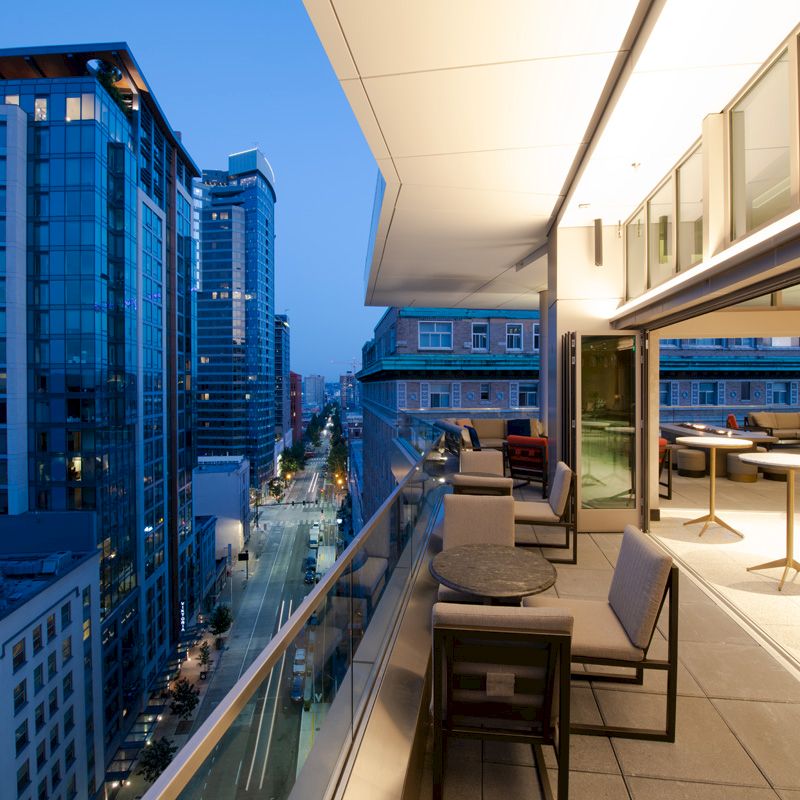 A modern cityscape at dusk with tall buildings, a well-lit balcony with tables and chairs, and a view of a street below.