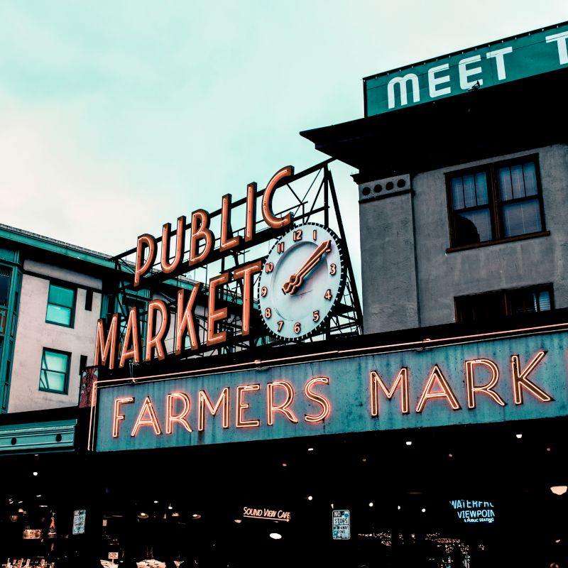 The image depicts a neon sign for a "Public Market" and "Farmers Market" with a clock, in an urban setting with surrounding buildings.