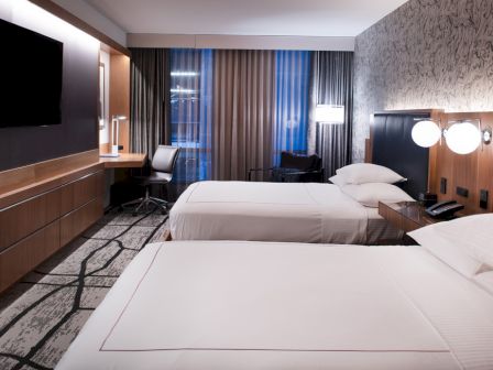 A modern hotel room with two double beds, stylish decor, a large flat-screen TV, a work desk, and large windows with curtains.