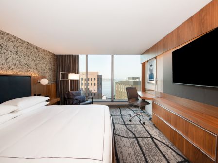 A modern hotel room with a large bed, floor-to-ceiling window, desk, chair, TV, and a pleasant city view.