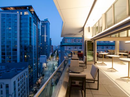 A modern cityscape with tall buildings is viewed from a balcony. The balcony has chairs, tables, and large windows, giving a sophisticated vibe.