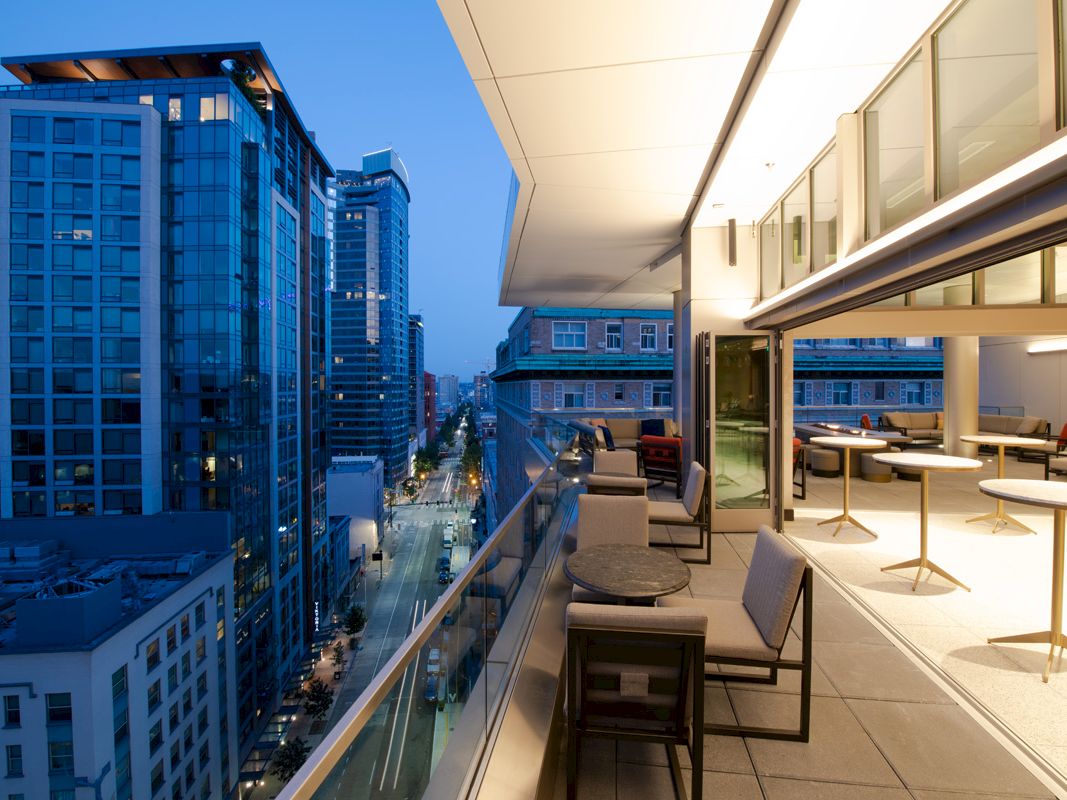 A modern cityscape at dusk featuring high-rise buildings and a spacious balcony with seating and tables, offering a blend of urban and elegant ambiance.