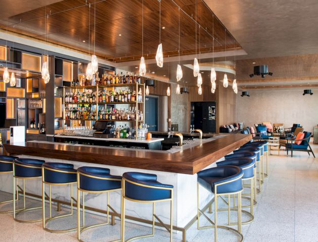 The image shows a modern bar with a well-stocked liquor shelf, bar stools, hanging lights, and a cozy seating area in the background.