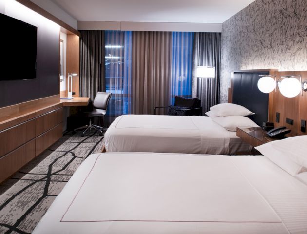 A modern hotel room features two neatly made beds, a wall-mounted TV, a desk with a chair, and contemporary lighting fixtures on the wall.
