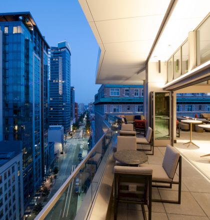 A modern balcony in a high-rise building at dusk overlooks a city street lined with other high-rise buildings, some with illuminated windows.