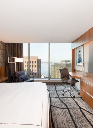A modern hotel room features a large bed, wall-mounted TV, desk, chair, and floor-to-ceiling windows with a city view, decorated in neutral tones.