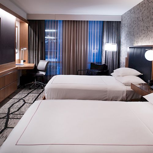 A modern hotel room with two beds, a desk and chair, a flat-screen TV, and contemporary lighting.