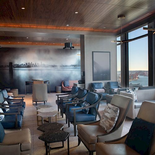 This image shows a modern lounge with plush armchairs, a wall mural, large windows offering a scenic water view, and a Ferris wheel in the distance.