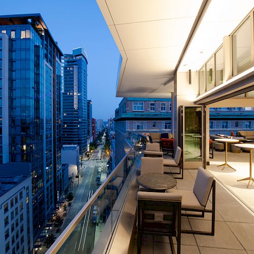This image shows a modern cityscape with tall buildings and a balcony, featuring outdoor seating and tables, captured during the evening.
