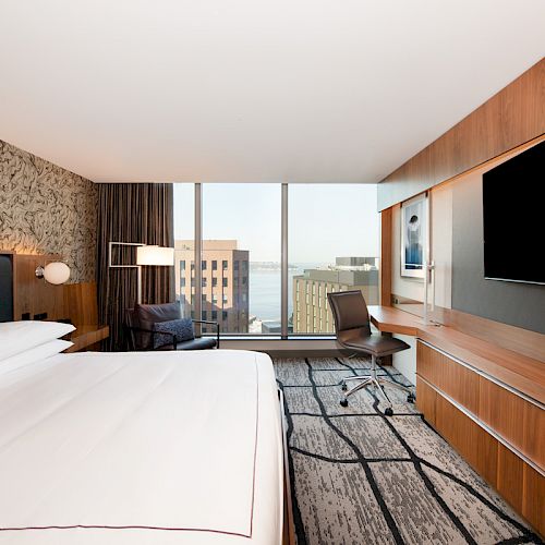 The image shows a modern hotel room with a large bed, a flat-screen TV, a work desk, a chair, and a window offering a cityscape view.