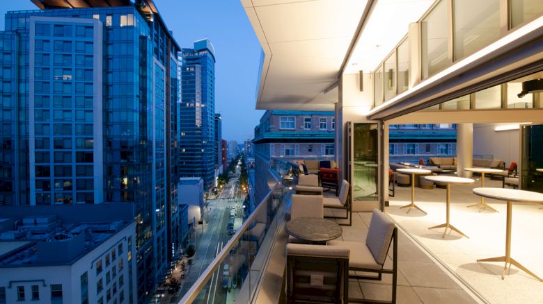 A modern terrace with outdoor seating overlooks a cityscape of tall buildings during twilight, providing a mix of urban and tranquil views.