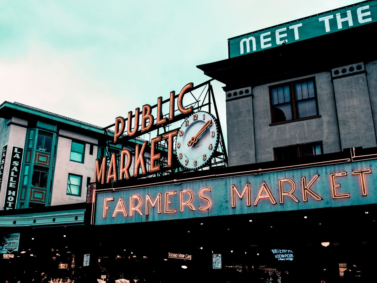 Image shows an outdoor view of a neon sign reading "Public Market" and "Farmers Market" with a clock, likely a popular city market area.