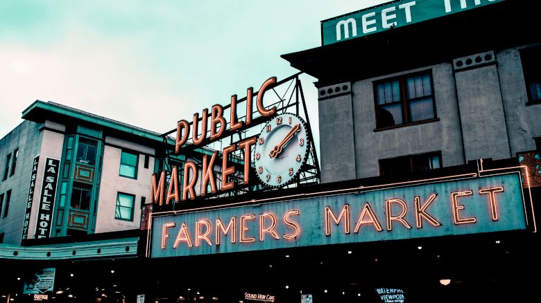Image shows an outdoor view of a neon sign reading "Public Market" and "Farmers Market" with a clock, likely a popular city market area.