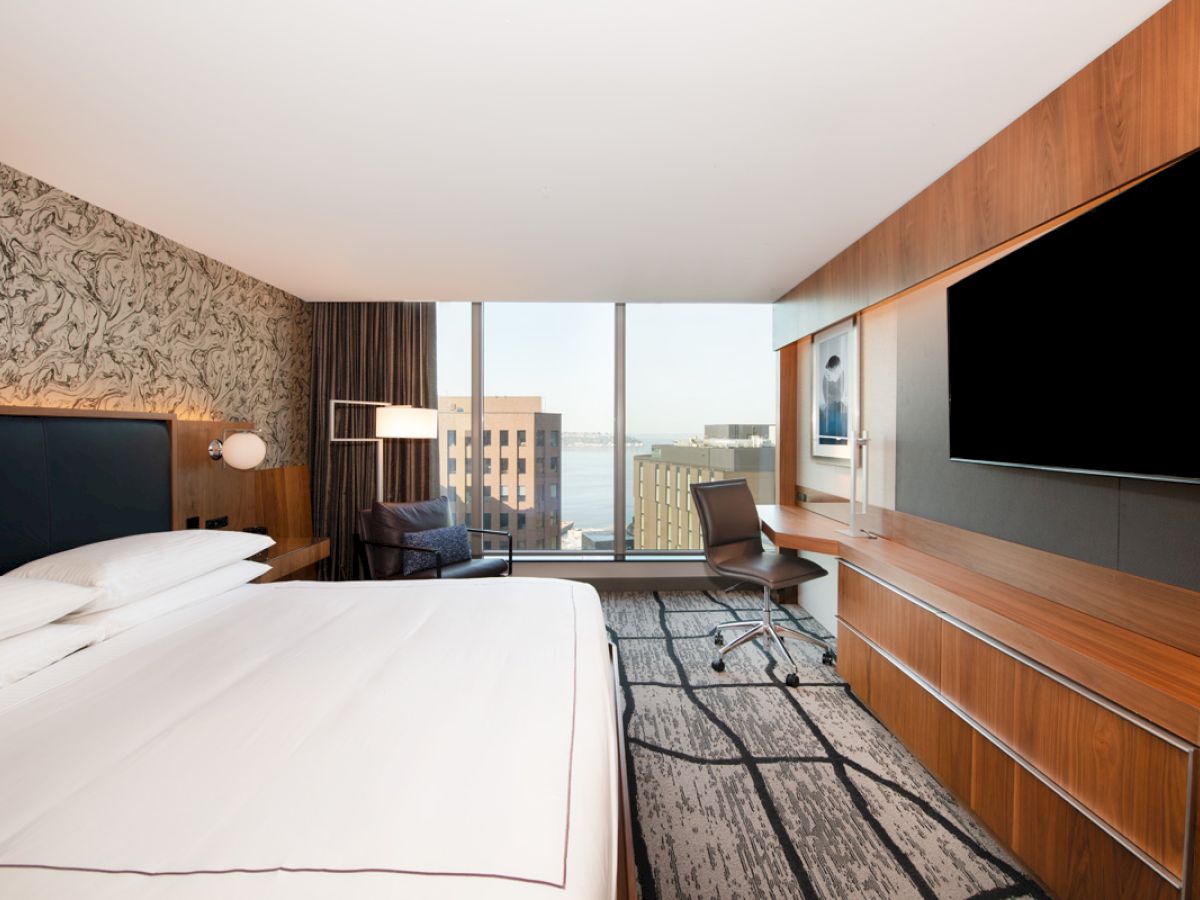 A modern hotel room with a large bed, desk, chair, television, and large window offering a city and water view, featuring minimalist decor.