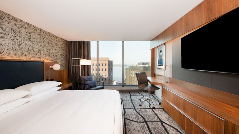 A modern hotel room with a large bed, desk, chair, television, and large window offering a city and water view, featuring minimalist decor.