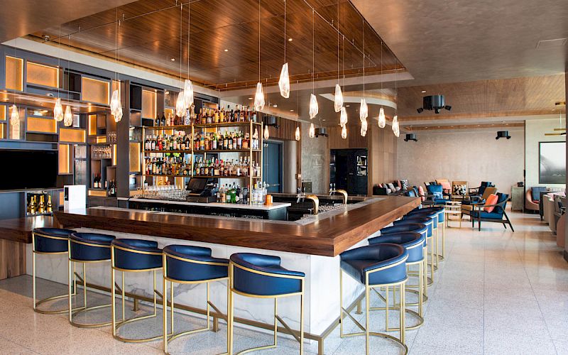 This image shows a modern bar with a sleek wooden counter, blue barstools, pendant lighting, and a fully stocked shelf of various bottles.