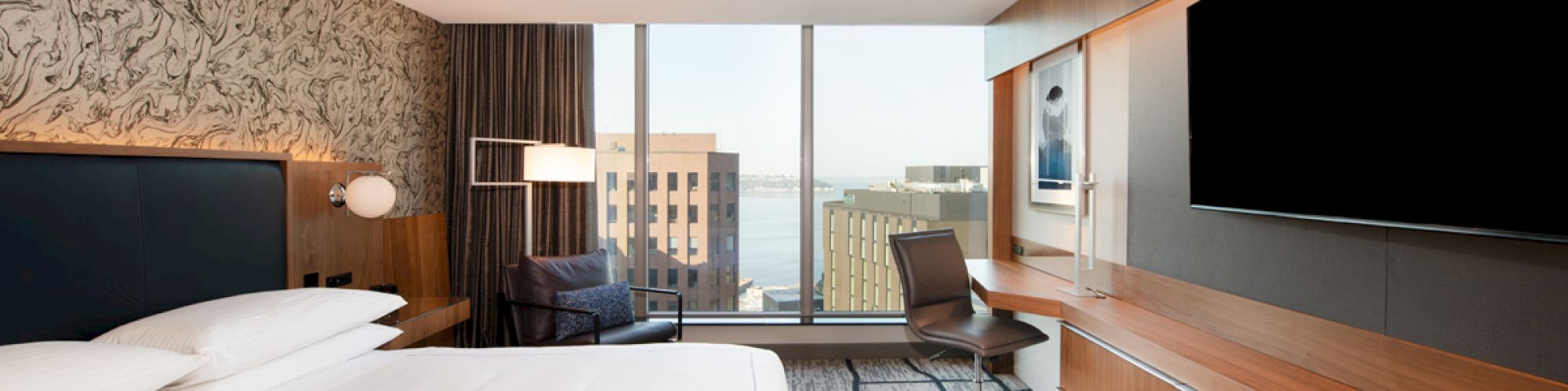 A modern hotel room features a bed, a desk, a chair, a large TV, and a view of buildings and water through a large window.